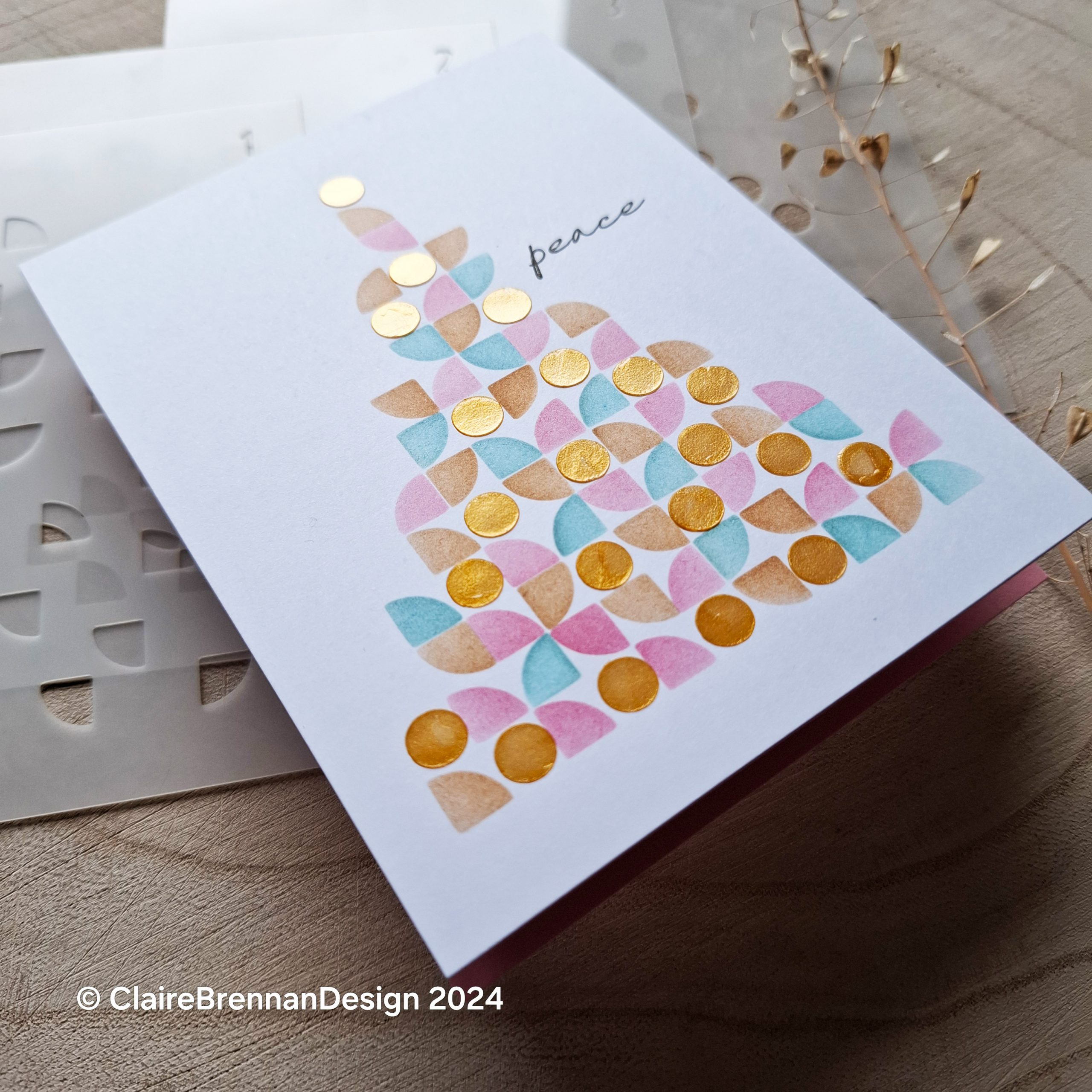 White Christmas card with abstract triangular Christmas tree shape, made of small geometric shapes, in pink and blue tones, on the front of a christmas card. The tipmost shape, and some of hte others is a shiny gold colour. The card rests on a neutral wooden background
