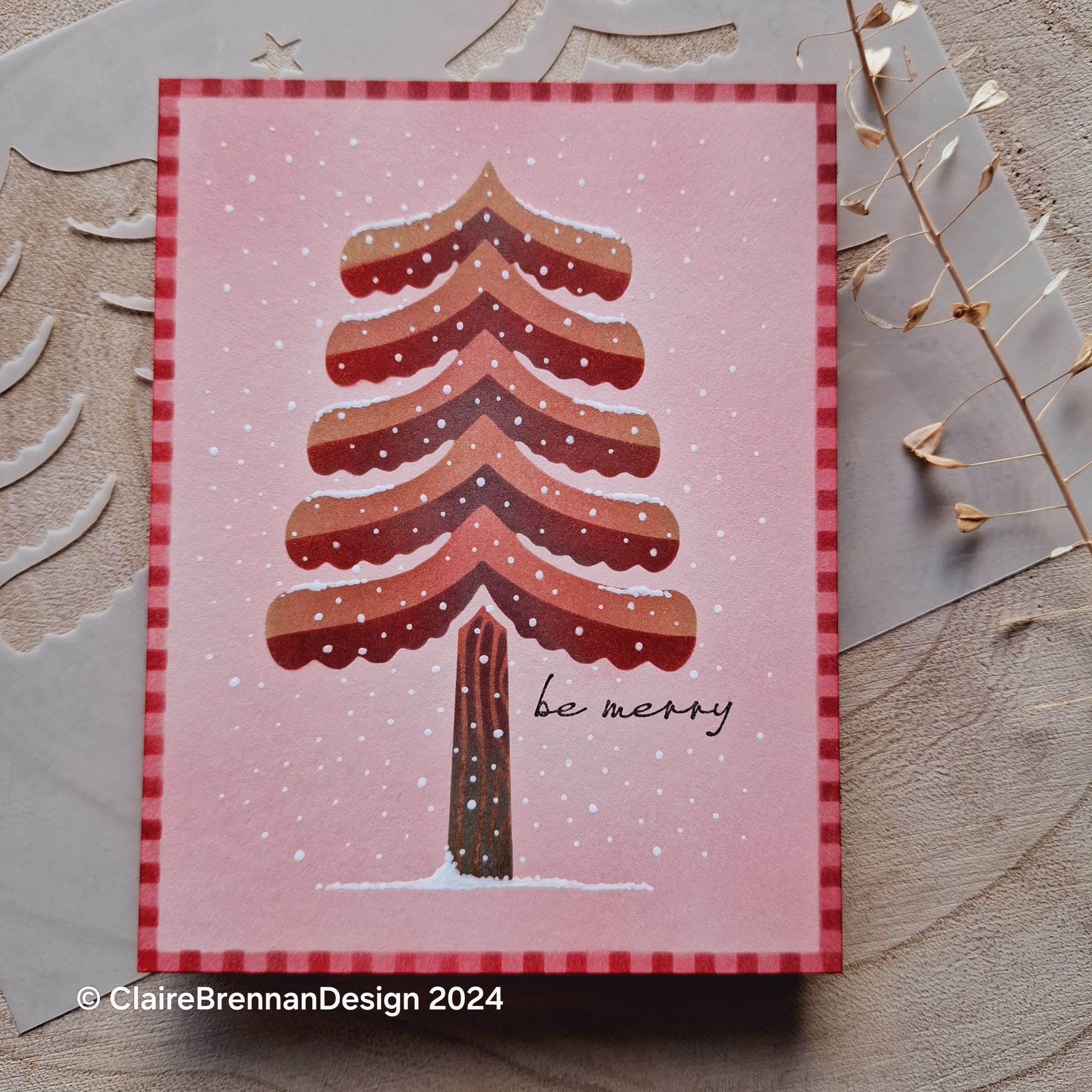 pink and red Christmas card featuring a folk art stylered and pink Christmas tree with white snow dots. card sits on a pale wooden tree trunk slice