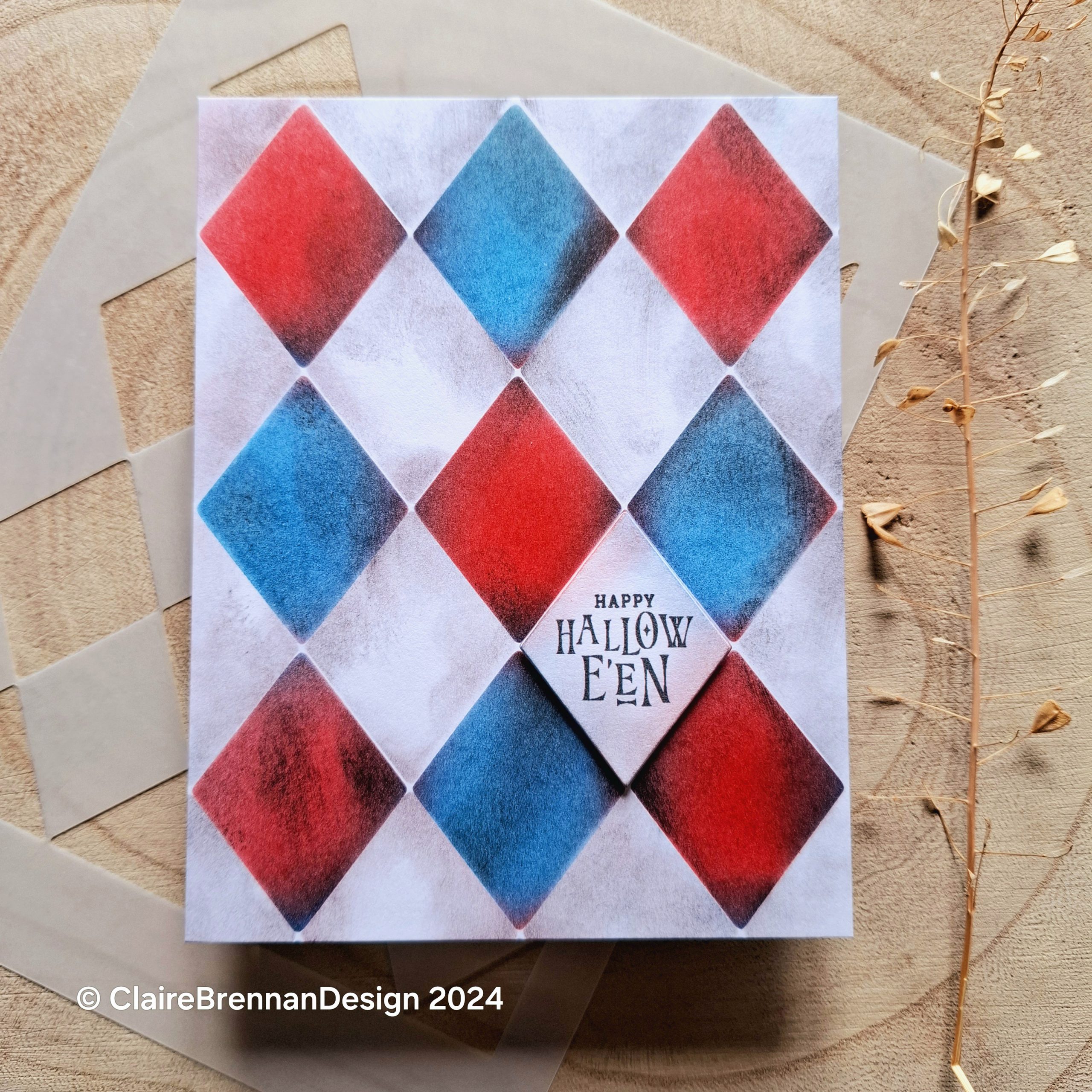 A Halloween greetings card with a red and white and blue grubby diamond harlequin pattern on the front, sitting on a slice of pale wooden tree trunk.
