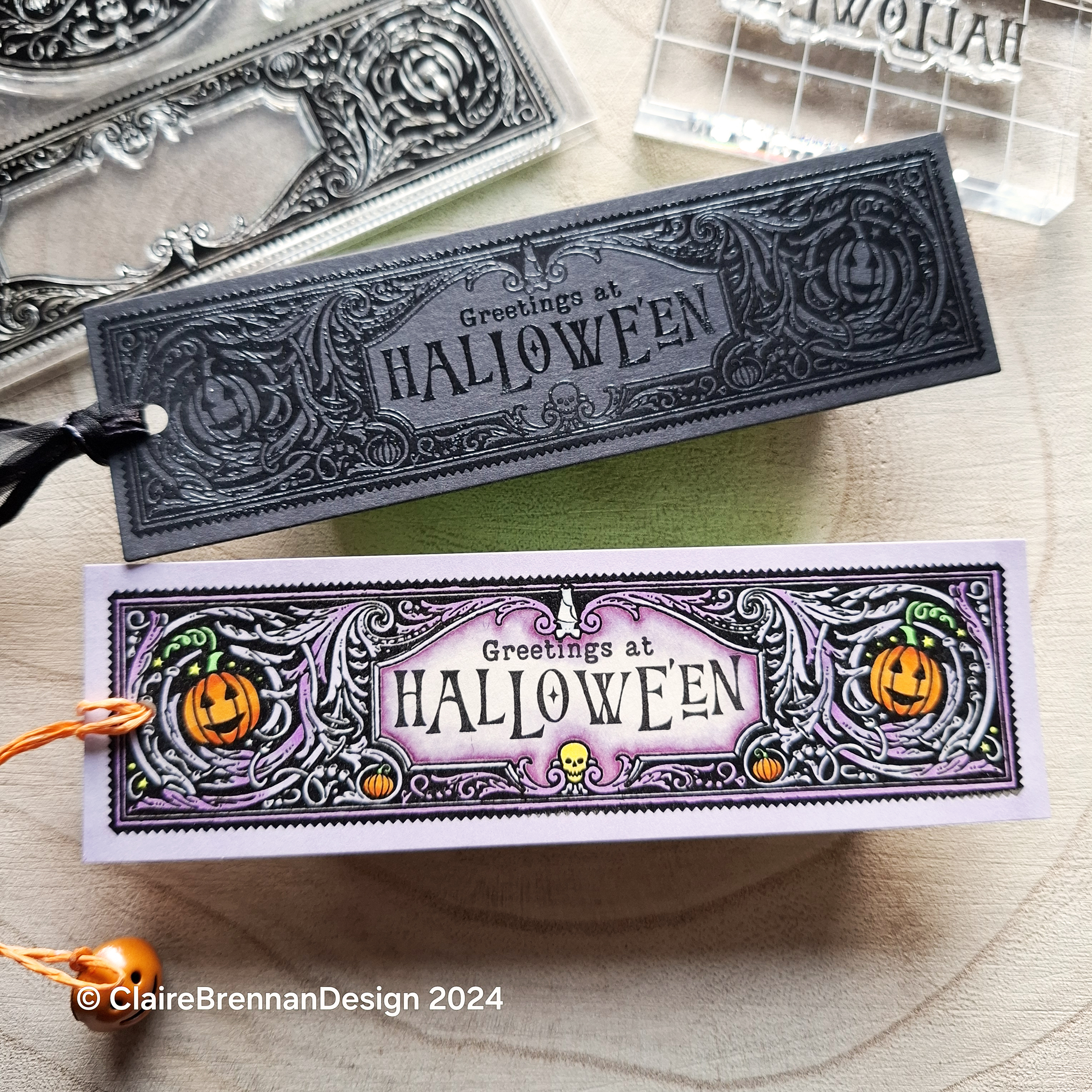 two bookmarks on a pale wooden background, one is black with black shiny pattern design, the other is pale purple with black orange and green Halloween design
