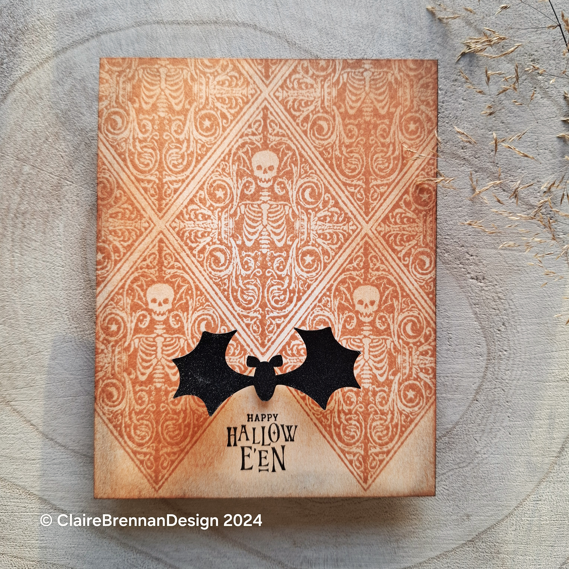 cream greeting card on wooden background, the card features a spooky diamond skeleton pattern stamped in orange ink, with a setiment and bat ornament in black.
