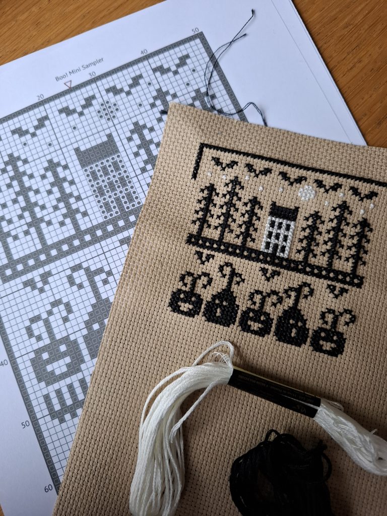 cross stitch in progress, black stitched trees beside a white stitched saltbox house