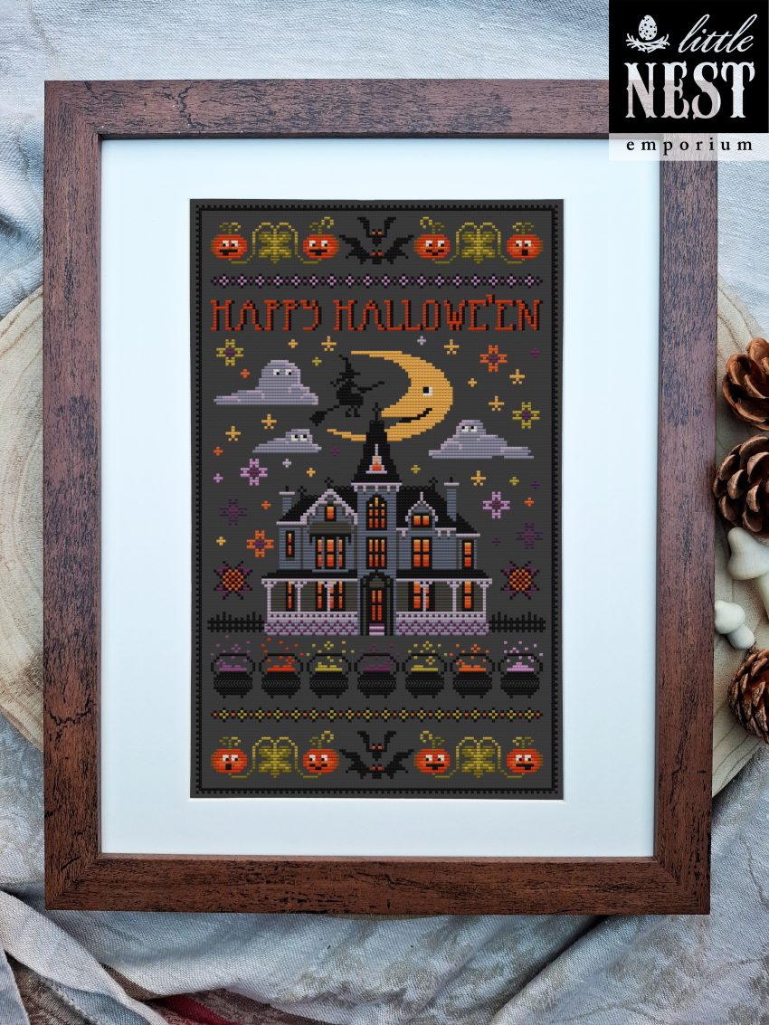 a dark grey piece of fabric with halloween images cross stitched on it in oranges and purples. The piece is framed in a a darkwood frame set on a pale wooden slice of tree trunk.