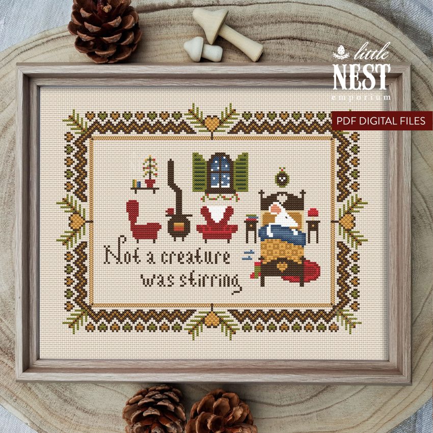 A Christmas themed cross stitch design on a cram background in a wooden frame - the cross sticth depicts a white mouse tucked up in a tiny wooden bed, in the background two red armchairs and a pot bellied stove.