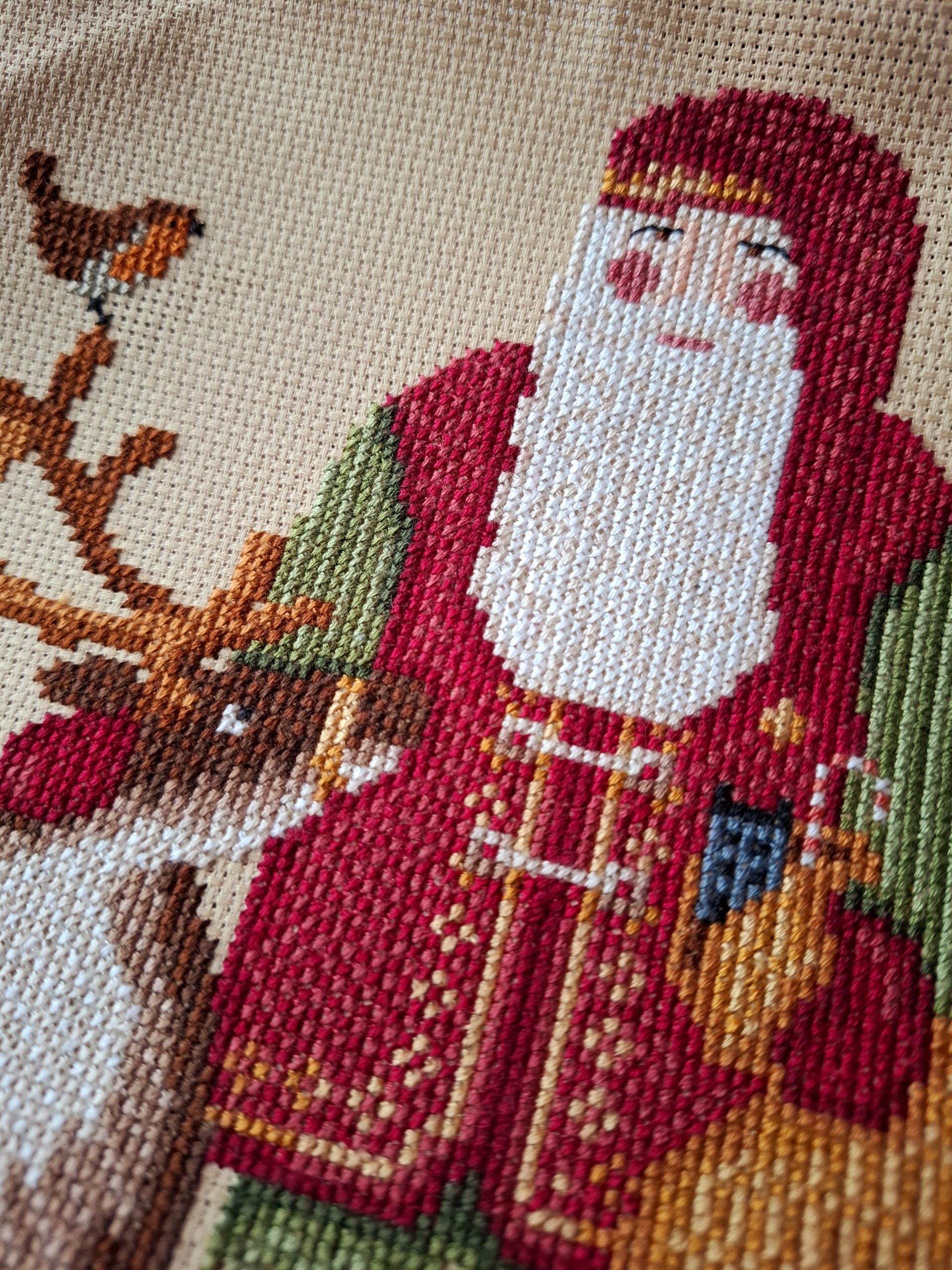 A cross stitch picture depicting a woodland santa figure in red waistcoat with green trousers. Santa pats a reindeer and a robin looks on